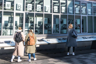 Drei Personen stehen vor einer Ausstellung mit dem Schrifzug "Besuch mich"