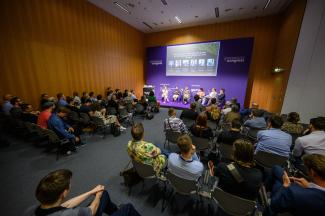 Blick in den Veranstaltungsraum, im Vordergrund sitzt das Publikum, im Hintergrund sind die Podiumteilnehmer zu sehen.